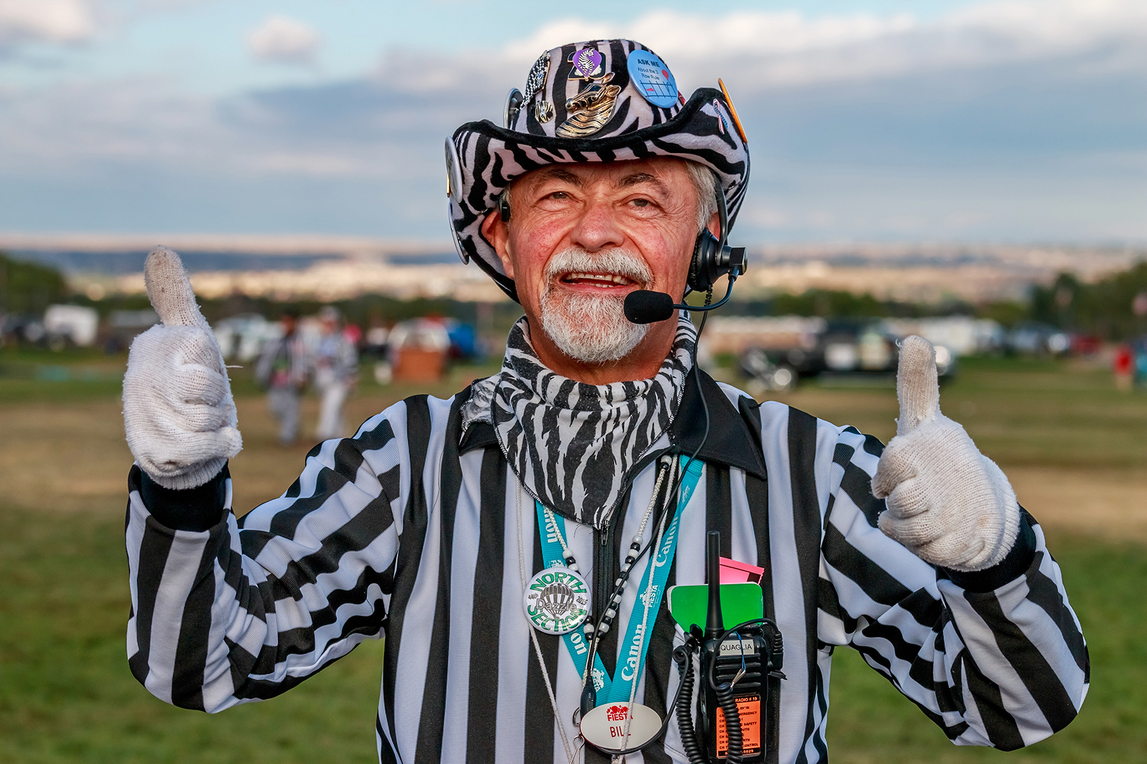 My 76/100 strangers is a fellow named Bill who described himself as intelligent, joyful and accommodating. Bill is a Zebra, and Chief Launch Director for the Albuquerque International Balloon Fiesta (AIBF). He has been involved with Fiesta for 21 years. He used to live in Albuquerque, but his wife’s job moved them away to Tennessee, where during the year he is a business to business consultant. Bill returns dutifully to Albuquerque to perform his mission critical AIBF event job, which shows his passion and commitment for the event. We met during Mass Ascension in an area that was being corded off for a hot air balloon compeition. Bill handed me a card while we were becoming acquainted. On one side is a group portrait of Zebras wearing their black and white dazzle uniforms. On the reverse it says the following: “The Launch Directors, affectionately known as the “Zebras” are volunteers whose job is to safely launch all 600+ balloons at the Albuquerque International Balloon Fiesta. The Zebras are all volunteers who work very hard for the nine days of Fiesta. Without their assistance, it would be close to impossible to get 600+ balloons in the air at the same time. They work hard, are dedicated and enjoy The Zebra life.” Like Bill, all Zebras dress in black and white striped referee uniform tops and each one puts custom flair touches on his or her outfit. Outlandish details are expected, such as zebra themed buttons or pins, and the zebra patterned cowboy hat you see being worn by Bill. Other than their microphone and earpieces for walkie-talkie radio communicaitions, Zebras all wear referee whistles and blow them to direct spectators and foot traffic away from balloon crews setting up the hot air balloon equipment, which includes balloon envelopes, stay lines and baskets. You usually hear them before you see them; which is hard to imagine considering the dazzle they must wear.